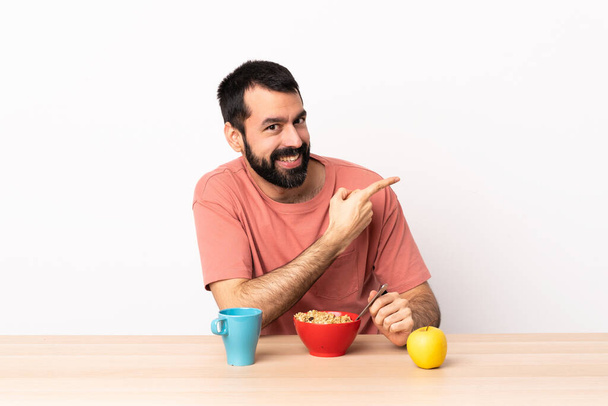 Mężczyzna rasy kaukaskiej jedzący śniadanie przy stole wskazującym na bok, aby przedstawić produkt. - Zdjęcie, obraz