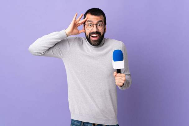 Uomo reporter adulto con barba che tiene un microfono su sfondo viola isolato con espressione a sorpresa - Foto, immagini