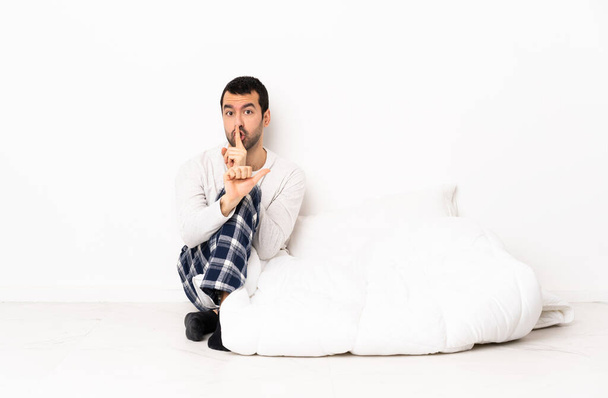 Blanke man in pyjama zittend op de vloer binnen wijzend naar de zijkant en het doen van stilte gebaar - Foto, afbeelding