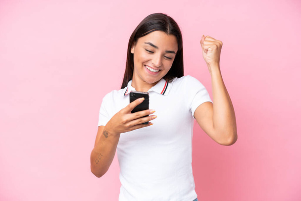 Giovane donna caucasica isolata su sfondo rosa utilizzando il telefono cellulare e facendo gesto di vittoria - Foto, immagini