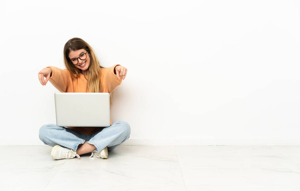 Jonge vrouw met een laptop zittend op de vloer wijzend vooraan met vrolijke uitdrukking - Foto, afbeelding