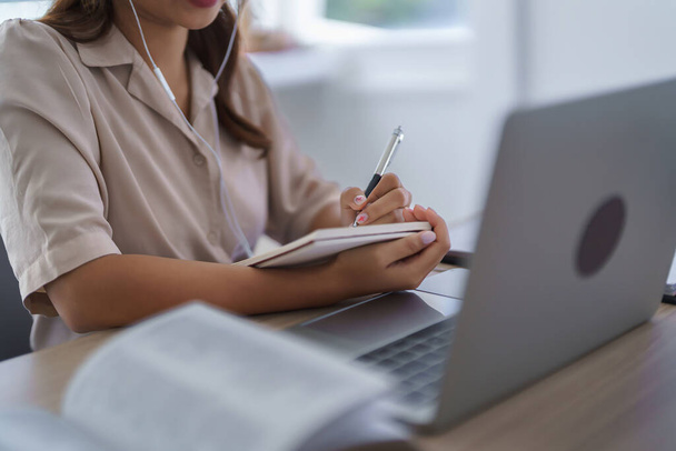 Młody azjatycki student w ubraniach casual pisanie danych wiedzy w notebooku podczas noszenia słuchawek do oglądania tutorial i studiuje edukację wykładowców klasy online z technologii laptopa w uniwersytecie. - Zdjęcie, obraz