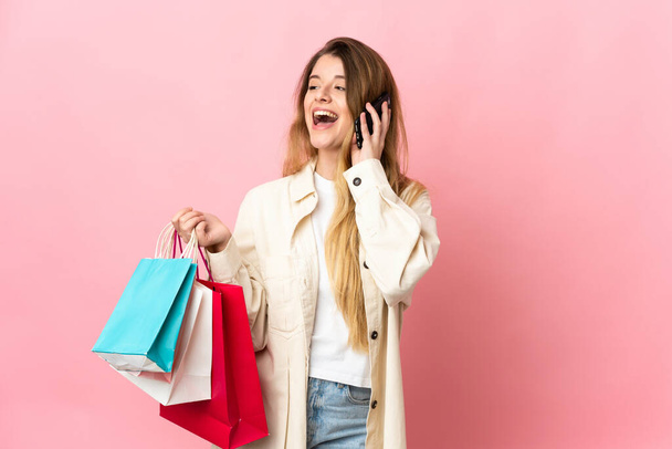 Fiatal nő bevásárló táska elszigetelt rózsaszín háttér gazdaság bevásárló táskák és hívja egy barátom a mobilját - Fotó, kép