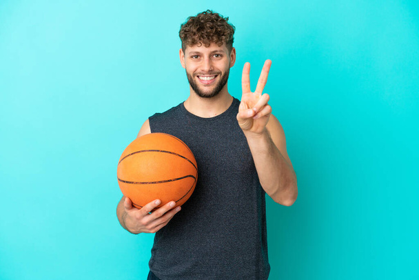 Bonito jovem jogando basquete isolado no fundo azul sorrindo e mostrando sinal de vitória - Foto, Imagem