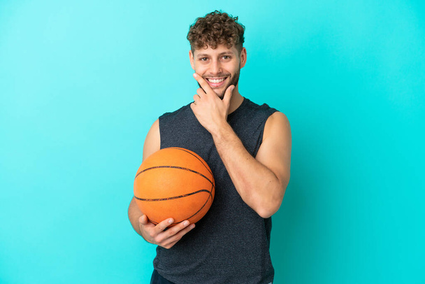Schöner junger Mann spielt Basketball isoliert auf blauem Hintergrund glücklich und lächelnd - Foto, Bild