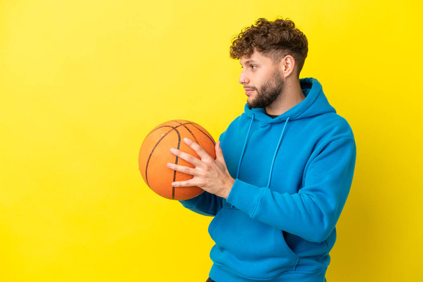 Jonge knappe blanke man geïsoleerd op gele achtergrond basketbal spelen - Foto, afbeelding