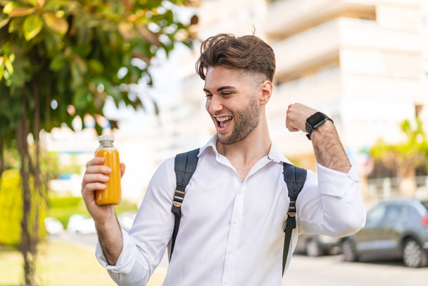 Young handsome man holding an orange juice at outdoors celebrating a victory - Photo, Image
