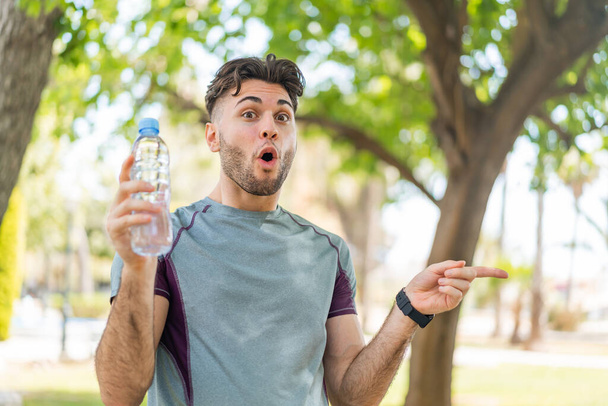 Jonge knappe sportman met een fles water buiten verrast en wijzend kant - Foto, afbeelding
