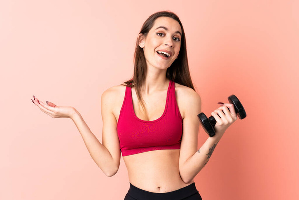 Jovem mulher esporte fazendo levantamento de peso sobre fundo rosa isolado com expressão facial chocada - Foto, Imagem