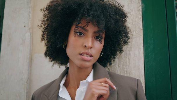Attractive pensive business woman looking camera standing at building wall closeup. Portrait of confident african american girl posing wearing stylish suit on street. Curly young lady standing outdoor - Photo, image