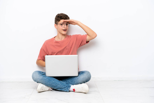 Adolescent russe homme tenant PC assis sur le sol isolé sur fond blanc regardant loin avec la main pour regarder quelque chose - Photo, image