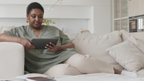 Donna nera che si riposa sul divano nel salotto moderno e naviga in Internet su tablet digitale durante il tempo libero a casa - Filmati, video