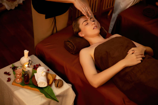 Kaukasische Frau genießt entspannende Anti-Stress-Kopfmassage und verwöhnt Schönheitspflege Gesichtshaut Freizeit in warmen Kerzenschein Ambiente Salon Spa in Luxus-Resort oder Hotel. Ruhige Lage - Foto, Bild