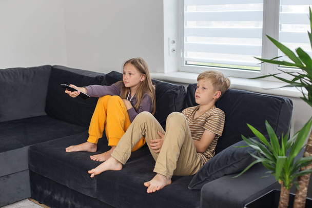 Παιδιά κάθονται στον καναπέ και βλέπουν τηλεόραση ενδιαφέρουσα ταινία. Το κορίτσι κρατάει τηλεκοντρόλ. Ξεκουράσου μετά το σχολείο. - Φωτογραφία, εικόνα