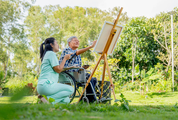 asiatico infermiera donna sedersi vicino anziano uomo su sedia a rotelle godere per dipingere il pittura all'aperto in il giardino e essi guarda felicità. - Foto, immagini