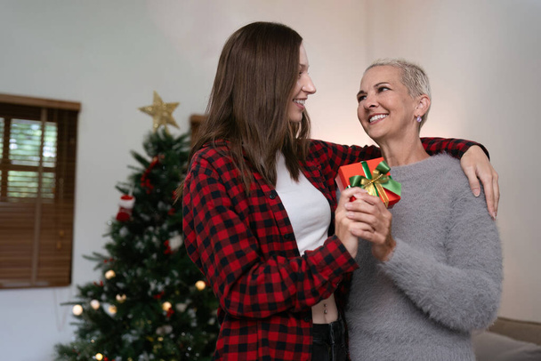 Fille câlin mère âgée avec boîte cadeau à la maison devant l'arbre décoré avec des décorations de Noël. - Photo, image