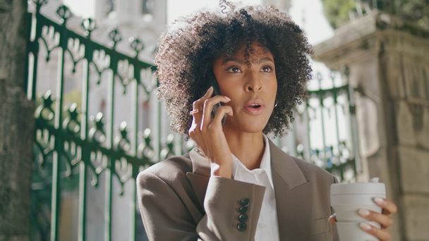 Πανέμορφη Αφροαμερικάνα που παίρνει τηλέφωνο κρατώντας καφέ απ 'έξω. Μια σγουρή νεαρή επιχειρηματίας που μιλάει στο τηλέφωνο στις σιδερένιες πύλες. Κομψό κορίτσι κρατώντας χάρτινο κύπελλο. - Φωτογραφία, εικόνα