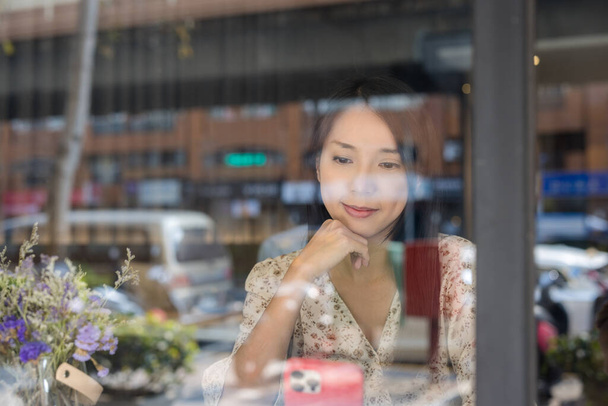 Kobieta korzystać z telefonu komórkowego i siedzieć wewnątrz kawiarni z odbiciem okna  - Zdjęcie, obraz