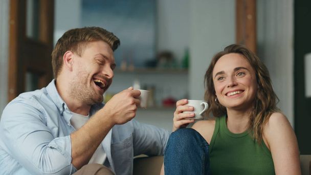 笑いの恋人コーヒーカップ自宅の近くに保持する。現代的な部屋でリラックスしたソファを楽しんでいる陽気な屈託のない配偶者。幸せな笑顔の妻夫共有面白い話飲みお茶で一緒に家 - 写真・画像