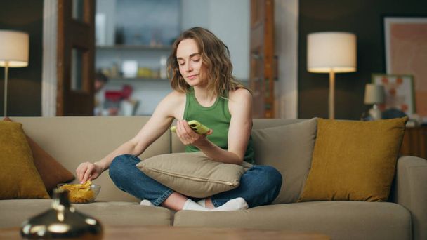 Relaxed girl eating snacks at cozy couch. Carefree woman unlocking mobile phone browsing social media at home. Calm unemployed lady spending lazy weekend at evening room interior. Free time concept - Photo, Image