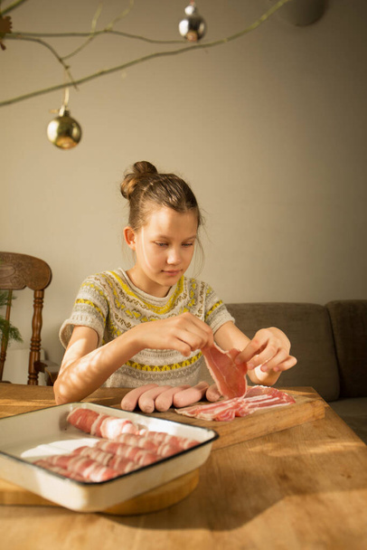 A girl prepares a traditional Christmas dish "Pigs in a blanket" Wraps sausages in bacon. Preparation for Christmas - Photo, Image
