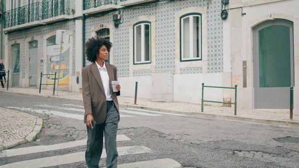 Sorrindo mulher atraente cruzando estrada da cidade segurando copo de papel de café takeaway. Curly afro-americana empresária caminhando passarela com smartphone moderno. Senhora feliz apressando-se em reunião de negócios. - Foto, Imagem