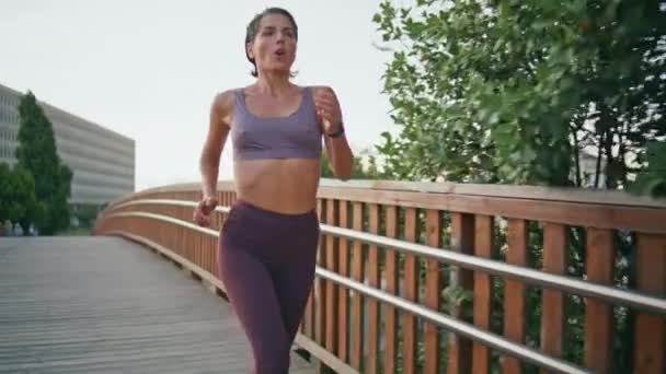 Nainen juoksee treenaamaan kardioharjoitusta puistossa. Urheilunainen juoksee sillalla. Fit urheilija lenkkeily yksin nauttia ulkona harjoitus. Motivoitunut urheilija kiiruhtaa vihreään luontoon. Terveiden elämäntapojen käsite - Materiaali, video