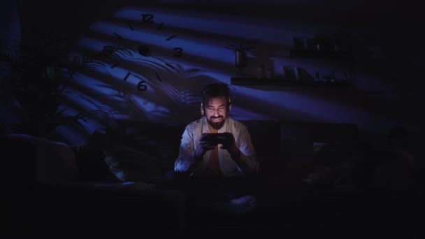Homme caucasien souriant regardant téléphone portable dans le salon avec écouteurs. Jeune garçon heureux d'utiliser ou de regarder des vidéos sur mobile assis sur le canapé la nuit. Concept d'applications de réseaux sociaux et de divertissement - Séquence, vidéo