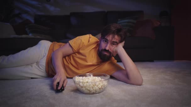 Νεαρός Ευρωπαίος με γενειάδα ξαπλωμένος στο χαλί του σαλονιού τρώγοντας ποπ κορν καθώς αλλάζει κανάλι χαμογελώντας ευτυχισμένος. Millennial τύπος απολαμβάνοντας ταινία ή σειρά και zapping με μπολ μέσα στο σπίτι τη νύχτα - Πλάνα, βίντεο