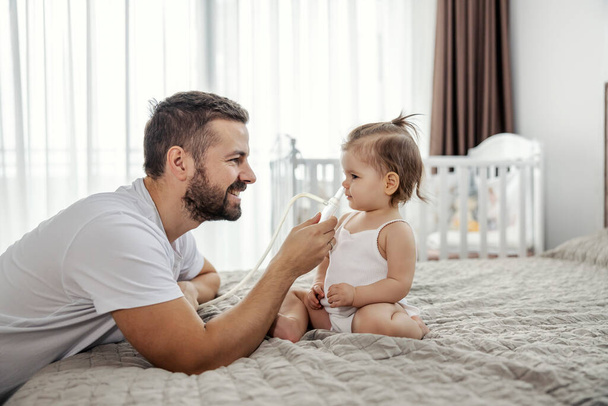 Ένας χαμογελαστός πατέρας καθαρίζει τη μύτη του κοριτσιού του με αντλία σε μια κρεβατοκάμαρα.. - Φωτογραφία, εικόνα
