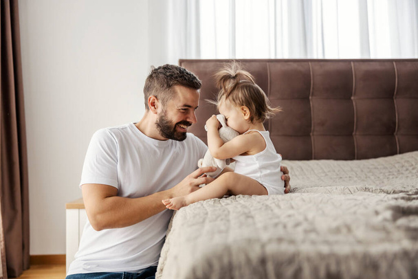 Un niño pequeño está sentado en una cama en un dormitorio y jugando con un juguete de conejo mientras su padre está sentado a su lado y riéndose de ella. - Foto, Imagen