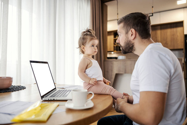 Un imprenditore occasionale è seduto a casa e parla con sua figlia mentre lavora a distanza su un computer portatile. - Foto, immagini