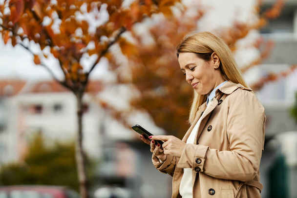 Une femme souriante échange des messages en ligne au téléphone en marchant dans la rue de la ville à l'heure d'automne. - Photo, image