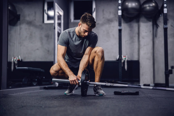 Working out in the gym with no other people, individual training. A shot of a handsome man in a grey shirt and shorts setting up equipment for training in a modern sports center. Fitness challenge - Photo, Image