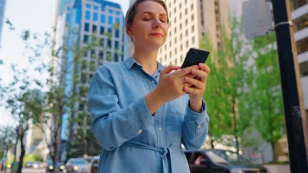 白人女性が街を歩き回り,スマートフォンを利用している. 背後には超高層ビルがいる. コミュニケーション,仕事の日,忙しい人生の概念. - 映像、動画