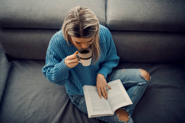 Een opgeleide vrouw nippen koffie tijdens het lezen van een interessant boek thuis. - Foto, afbeelding