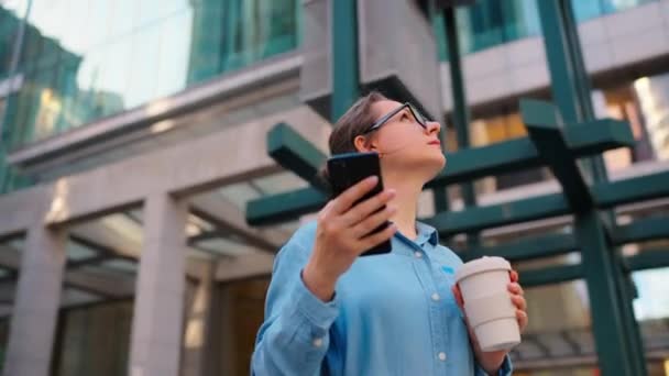 Белая женщина в очках стоит на улице, пользуется смартфоном и пьет кофе. Орбитальный выстрел. Небоскрёбы на заднем плане. Работа, рабочий день, занятость.. - Кадры, видео