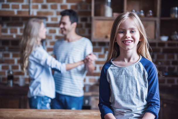 Cute little girl is looking at camera and smiling while her beautiful parents are dancing in the background in kitchen - Photo, Image