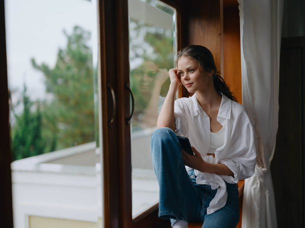 Νεαρή γυναίκα κάθεται σε ένα ξύλινο περβάζι παράθυρο με ένα τηλέφωνο στο χέρι χαμογελώντας και κοιτάζοντας έξω από το παράθυρο, φθινοπωρινή διάθεση, ζεστή ατμόσφαιρα στο σπίτι, αισθητικό τρόπο ζωής. Υψηλής ποιότητας φωτογραφία - Φωτογραφία, εικόνα