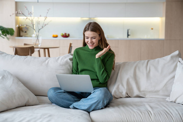 快適なソファーに座って快適な陽気な女性を微笑み,膝の上にラップトップコンピュータ,海外に住んでいる親戚とのオンラインビデオ通話,ボーイフレンド,友達とチャット  - 写真・画像
