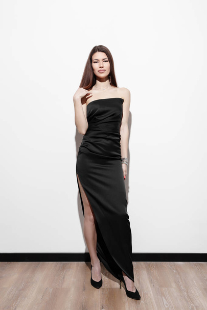 Kobieta stojąca pewnie w eleganckiej czarnej sukni wieczorowej bez ramiączek, jej subtelny uśmiech niosący ponadczasową łaskę - Zdjęcie, obraz