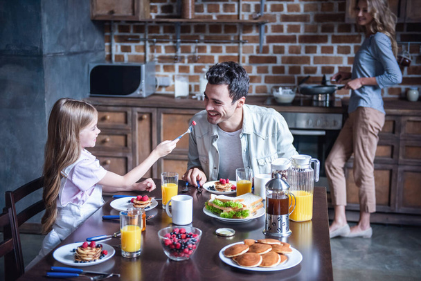 Onnellinen perhe aamiaisella keittiössä. Isä ja tytär istuvat pöydässä, kun äiti tekee pannukakkuja.
 - Valokuva, kuva