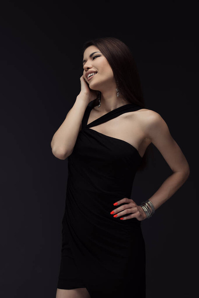 Une femme joyeuse dans une élégante robe noire à une épaule rit en posant, son bonheur accentué par un vernis à ongles rouge frappant et des bijoux sophistiqués - Photo, image