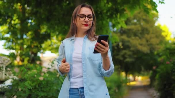 Gelukkige vrouw met laptop en smartphone in haar handen wandelen door de bloeiende tuin of groene straat na het werk dag. Langzame beweging - Video