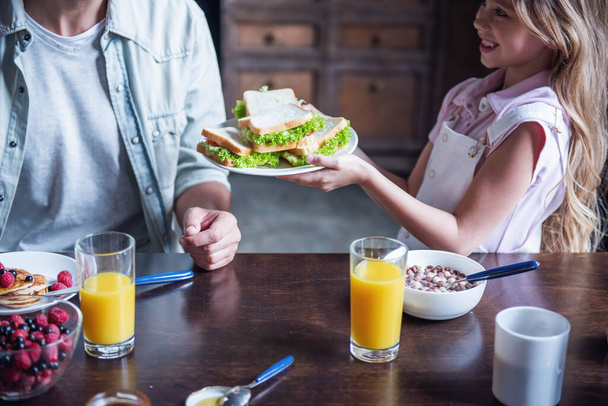 Happy family having breakfast in kitchen. Little girl is bringing sandwiches to her parents and smiling - Photo, Image