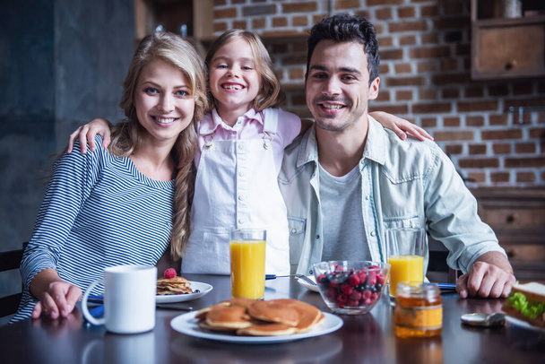 Onnellinen perhe halailee, katselee kameraa ja hymyilee syödessään aamiaista yhdessä keittiössä
 - Valokuva, kuva