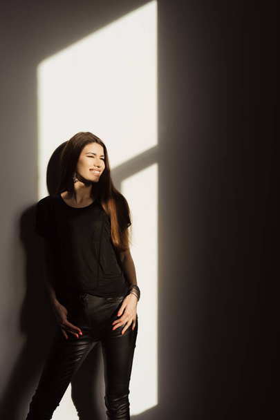 Una donna in abbigliamento casual nero sorride mentre gode del calore della luce solare naturale che scorre attraverso una finestra, creando un suggestivo gioco di luci e ombre. - Foto, immagini