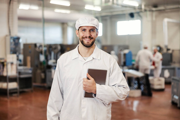 Un superviseur d'usine de viande heureux tient la tablette dans ses mains et se prépare à évaluer la qualité de la nourriture tout en se tenant debout dans l'installation et en souriant à la caméra. - Photo, image