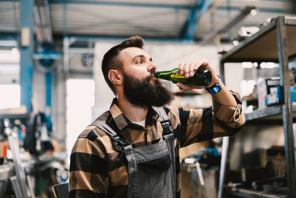 Un operaio che beve birra in fabbrica durante una pausa. - Foto, immagini