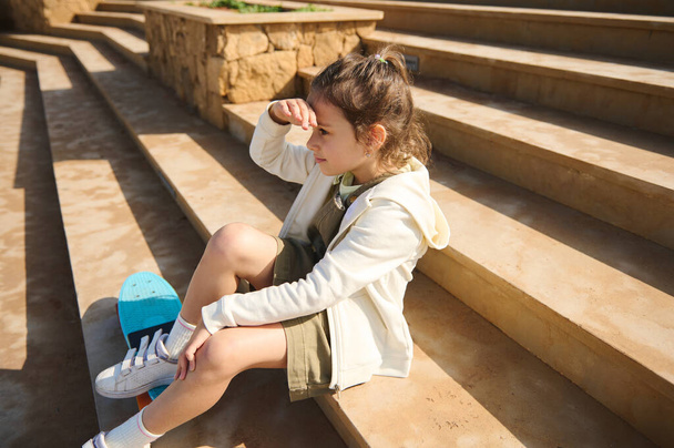 Imádnivaló kaukázusi óvodás lány néz a távolba, ül a szabadban a lépcsőn, miután pihenés után gördeszka egy játszótéren, öltözött bézs kapucnis pulcsi és khaki ruha - Fotó, kép